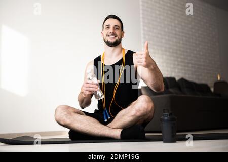 Giovane uomo che si esercita a casa. Guy si siede da solo sulla mappa dello yoga nero, tiene una bottiglia d'acqua e pollici. Corda gialla che salta intorno al collo. Athletist riposo e. Foto Stock