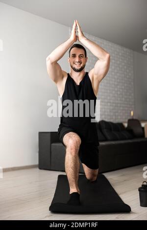 Uomo che pratica yoga indoor facendo Warrior uno su tappetino nero. Giovane muscoloso che si rilassa e si equilibra in asana. Esercizio mattutino. Mente sana in s. Foto Stock