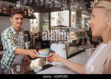 Cordiale e simpatico barista maschile che serve una tazza di caffè per la sua cliente femminile. Giovane allegro che si diverte a lavorare al suo bar, dando una tazza o Foto Stock