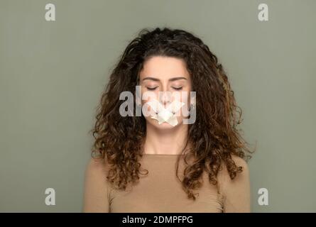 Giovane donna con gli occhi chiusi e la bocca chiusa in un concetto di censura della visione e della libertà di parola su uno sfondo grigio dello studio Foto Stock