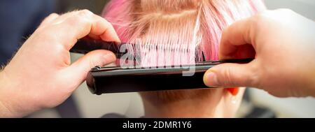 Primo piano vista posteriore delle mani del parrucchiere che raddrizzano il rosa corto capelli con una piastra per capelli in un parrucchiere Foto Stock