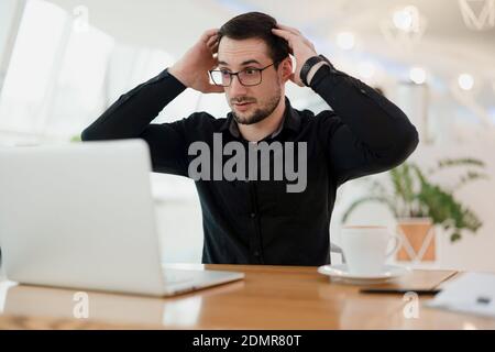 Che cosa sta accadendo uomo intelligente scioccato in occhiali guardando sul suo laptop e tenendo la testa per le mani. Errore nel programma. Il programmatore maschile è scioccato Foto Stock