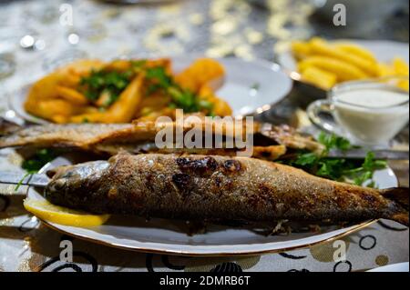 Close up of fried trout on white dish with golden border with lemon, garnish and sauce in the background. Selective focus Foto Stock