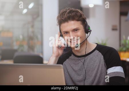 Allegro bel giovanotto sorridente alla telecamera, che lavora al call center, spazio per la copia. Attraente operatore di assistenza clienti maschile indossando cuffie w Foto Stock