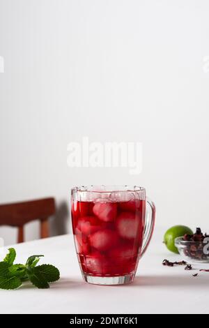 Tè hibiscus freddo rinfrescante in un bicchiere di ghiaccio, foglie di tè secco di tisana rossa con calce sullo sfondo e menta aromatica fresca Foto Stock