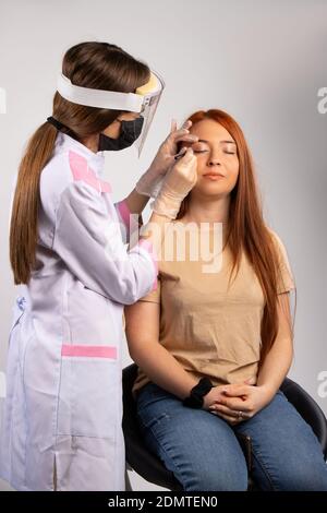 Un make-up artista in un abito medico, maschera protettiva e guanti facendo un trucco per una ragazza con capelli rossi. Servizio di bellezza in concetto pandemico. Foto Stock