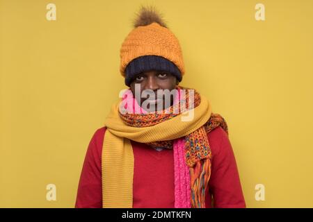 Divertente uomo afroamericano maturo in sciarpa gialla e cappello che si gelano in abiti caldi. Riprese in studio su parete blu. Foto Stock