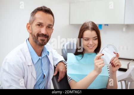 Bel medico maschio maturo sorridendo alla telecamera, la sua paziente femmina sorridente guardando nello specchio. Giovane donna che controlla i suoi denti nello specchio afte Foto Stock