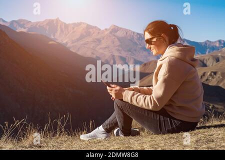 Giovane donna con telefono si siede a terra contro lo sfondo di montagne, luce solare Foto Stock