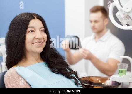 Controlli orali. Bella donna anziana sorridente guardando via seduta in una sedia dentale presso la clinica il suo dentista che esamina i denti x-ray sullo sfondo Foto Stock