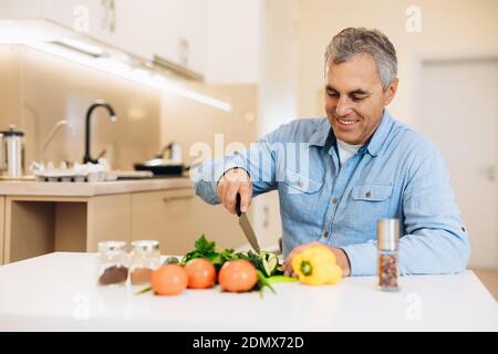 Un vecchio uomo allegro taglia le verdure con un coltello in una cucina con mobili bianchi. Mangia più verdure spezie e verdure sfocate in primo piano. Gustoso v Foto Stock