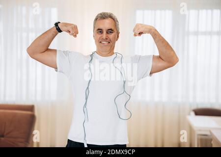 Allegro uomo maturo mostrando i suoi bicipiti e sorridendo. Concetto di allenamento duro. Soggiorno luminoso sullo sfondo. Uomo adulto che mostra i muscoli. Blu Foto Stock