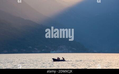 Perast, Cattaro, Montenegro. Vista attraverso la baia di Cattaro, sera, piccola barca dwarfed dal paesaggio di alta montagna. Foto Stock