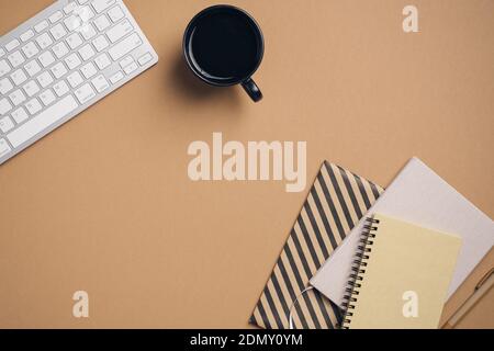 Moderna scrivania da ufficio con tastiera, tazza di caffè, taccuini e blocco note su sfondo marrone pastello. Elegante spazio di lavoro per freelance. Business concep Foto Stock