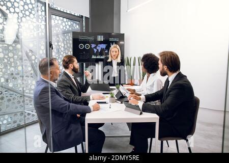 Consiglio di amministrazione di diversi dirigenti multietnici uomini d'affari durante la riunione annuale in sala d'ufficio, ascoltando matura signora bionda e discutendo i risultati del lavoro. TV Foto Stock