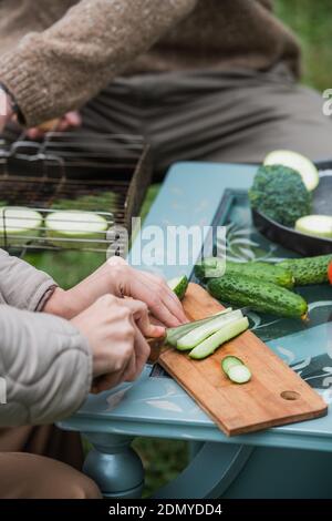 Colpo di fermo, mani che tagliano le verdure su un tagliere. Preparandosi per una cena di famiglia sul prato di fronte ad una casa di campagna. Attività estive Foto Stock
