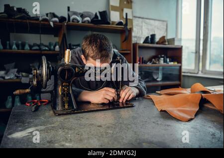 A young shoemaker in his workshop sews leather on a sewing machine, makes original shoes to order. Stock Photo