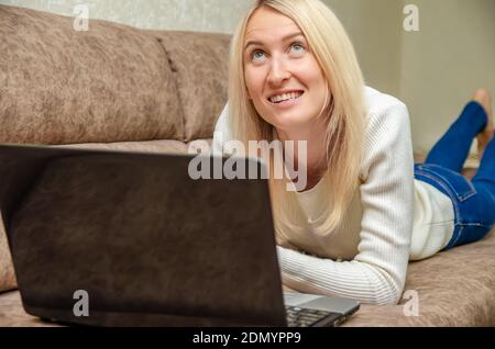 Young blonde woman is lying on the sofa with a laptop, smiling and looking up mysteriously. Concept of online communication, self-isolation Foto Stock