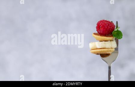 Forchetta da primo piano con minuscoli pancake, fetta di banana, lampone e foglia di menta. Spazio per il testo, messa a fuoco selettiva Foto Stock