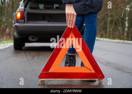 giovane donna in una giacca invernale in un giallo vest mette un segnale di arresto di emergenza sulla strada an triangolo arancione vicino a un'auto con un'auto aperta trunk Foto Stock