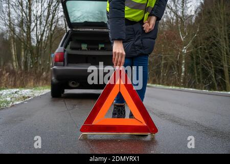 giovane donna in una giacca invernale in un giallo vest mette un segnale di arresto di emergenza sulla strada an triangolo arancione vicino a un'auto con un'auto aperta trunk Foto Stock