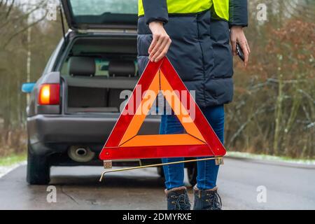 giovane donna in una giacca invernale in un giallo vest mette un segnale di arresto di emergenza sulla strada an triangolo arancione vicino a un'auto con un'auto aperta trunk Foto Stock