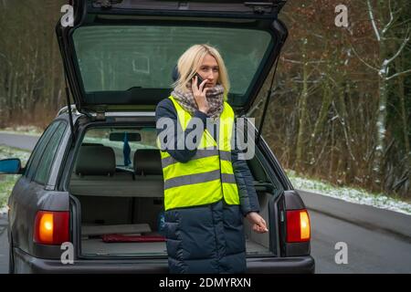 Giovane donna bionda in una giacca invernale in un giubbotto giallo è in piedi vicino a un'auto con un aperto trunk e conversazione al telefono Foto Stock