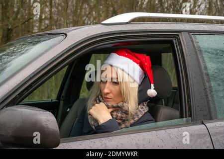 Giovane bionda donna in un cappello di Babbo Natale è seduta al volante di una macchina e guarda con un sguardo attento e triste in lontananza Foto Stock