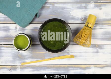 Vista dall'alto su un tavolo di legno bianco con ciotola di sciawano, bambu whisk e cucchiaio, tisana verde lattina e tovagliolo blu Foto Stock