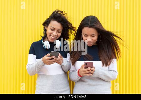 Two smiling brunette girls on a yellow background. They are wearing the same dress and are using their smart phones. Space for text. Stock Photo