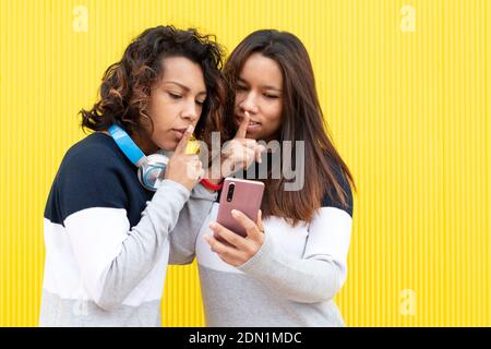 Ritratto di due ragazze marroni su sfondo giallo. Entrambi fanno il gesto di chiedere il silenzio mentre si guarda uno smartphone. Spazio per il testo. Foto Stock