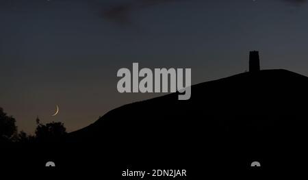 Una nuova Luna ambientata che scende dietro Glastonbury Tor, Somerset, Inghilterra, Regno Unito, in una serata estiva. Foto Stock