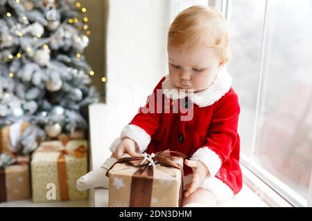 Ragazza piccola graziosa rossa in vestito rosso che gioca con natale confezione regalo mentre si è seduti sul davanzale Foto Stock