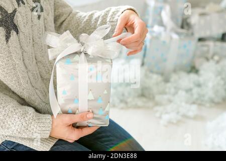 Close-up female hands tie the white bow of a gift box with silver wrapping. Christmas crafts. Stock Photo