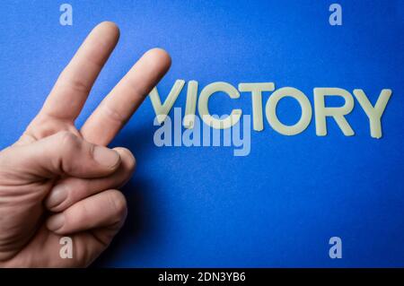 Mano umana che fa un segno di vittoria accanto alla parola scritta con lettere di plastica su sfondo di carta blu, concetto Foto Stock