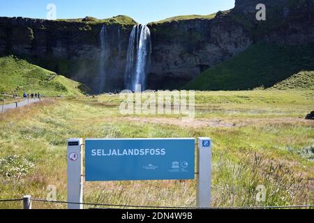 Un sentiero conduce dal parcheggio verso la cascata Seljalandsfoss in Islanda del Sud, appena fuori dalla circonvallazione. Un cartello blu si trova accanto al passaggio pedonale. Foto Stock
