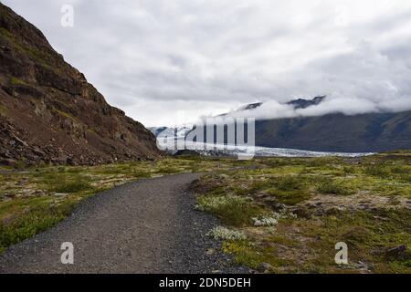 Un sentiero lungo un sentiero in ghiaia che si dirige verso il ghiacciaio Skaftafellsjokull a Skaftafell, Parco Nazionale Vatnajokull, Islanda. Nuvole basse nella valle. Foto Stock