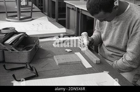 1964, storico, uno studente di architettura maschile dell'Università seduto a una scrivania usando una pentola di colla per unire una carta per fare un modello, California, USA. Foto Stock