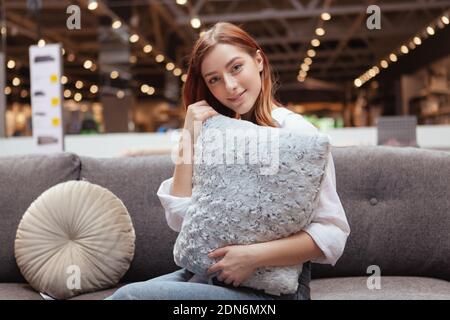 Bella donna allegra abbracciando un cuscino, seduta su un nuovo divano al negozio di mobili Foto Stock