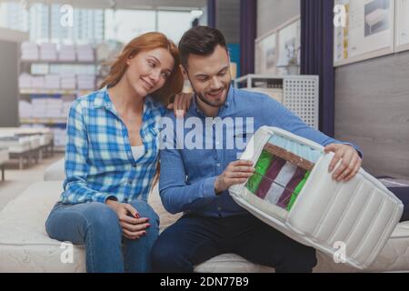 Bell'uomo allegro che si diverte a fare acquisti di materassi con la sua bella moglie. Una coppia affascinante che sceglie un nuovo letto ortopedico nel negozio di mobili. Felice co Foto Stock