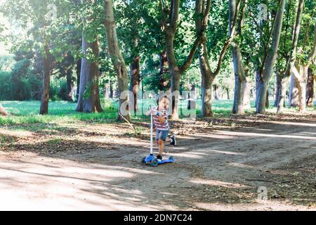 Bambino su scooter nel parco. I bambini imparano a skate roller board. Piccolo ragazzo che pattina nella soleggiata giornata estiva. Attività all'aperto per bambini in strada sicura. Foto Stock