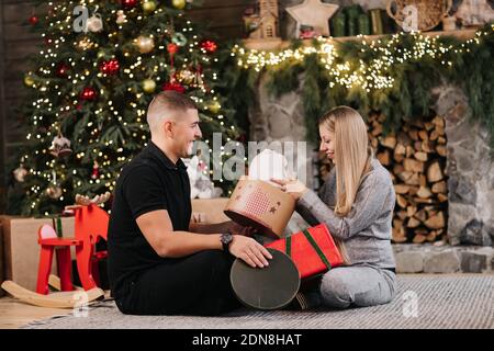 Adorabile coppia di matrimonio sedersi vicino albero di Natale e camino a casa, marito cura abbraccio bella moglie incinta, giovane famiglia godere di felicità, wint Foto Stock