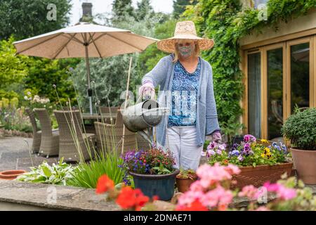 Donna innaffiatura piante e fiori in giardino con una maschera di copertura durante il Coronavirus Covid-19 pandemia in blocco Foto Stock
