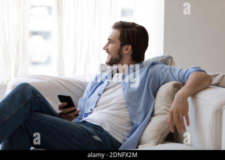 Felice giovane uomo che si rilassa sul divano, tenendo lo smartphone nelle mani. Foto Stock