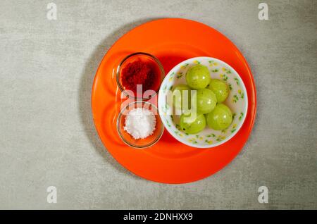 Amla crudo, uva spina indiana, emblica di Phyllanthus. Acidulo a piacere con polvere di peperoncino rosso, sale in piatto arancione su sfondo grigio astratto Foto Stock