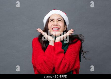 Sorpresa eccitato bella donna asiatica in rosso maglione di Natale e. cappello sorridente con mani aperte sotto il mento studio girato sopra sfondo grigio isolato Foto Stock