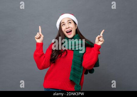 Donna asiatica felice in rosso e verde abbigliamento di Natale cercando e poiting mani in su in grigio isolato studio sfondo Foto Stock