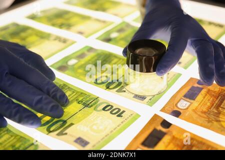 Mani in guanti di gomma che tengono la lente d'ingrandimento sopra le banconote closeup Foto Stock