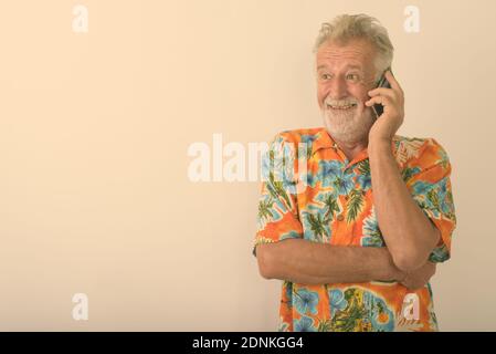 Studio shot di felice senior turistico barbuto uomo sorridente e pensare mentre parlano al telefono mobile contro uno sfondo bianco Foto Stock