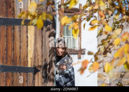 Una ragazza sorridente con un gatto bianco guarda fuori da dietro una porta di fienile rustico in legno in un giardino d'autunno Foto Stock